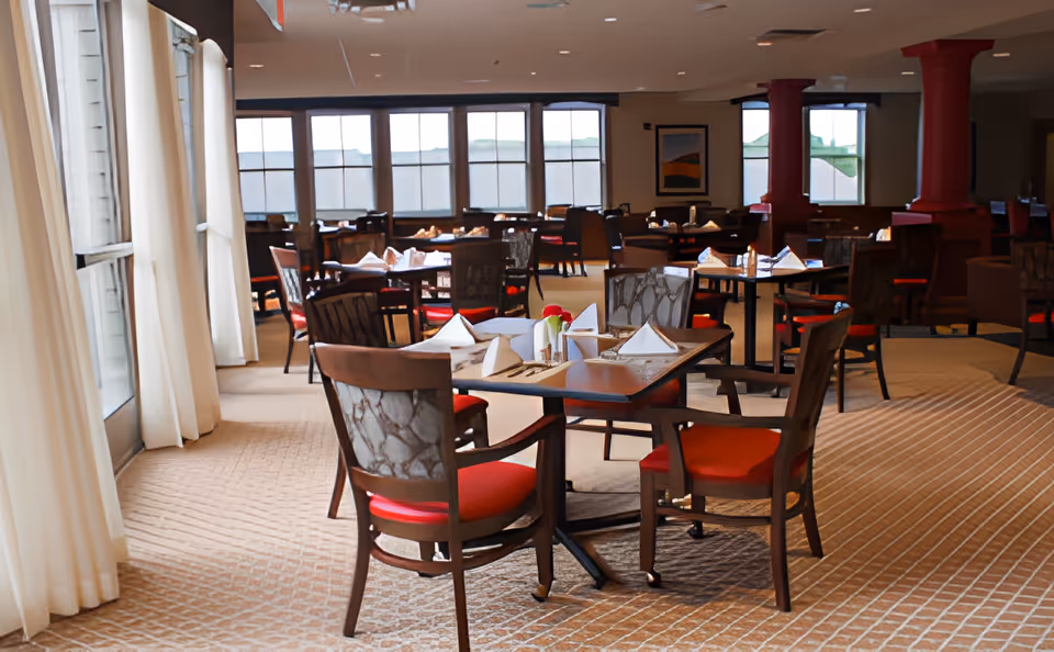 A spacious dining room in The Pines Senior Living with multiple tables set with white napkins, cutlery, and small flower vases. The room features large windows with sheer curtains allowing natural light to fill the space. Chairs with red cushions surround the tables, and the carpet has a patterned design.