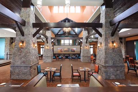 Spacious dining room with stone columns, exposed wooden beams, chandeliers, and arranged tables and chairs.