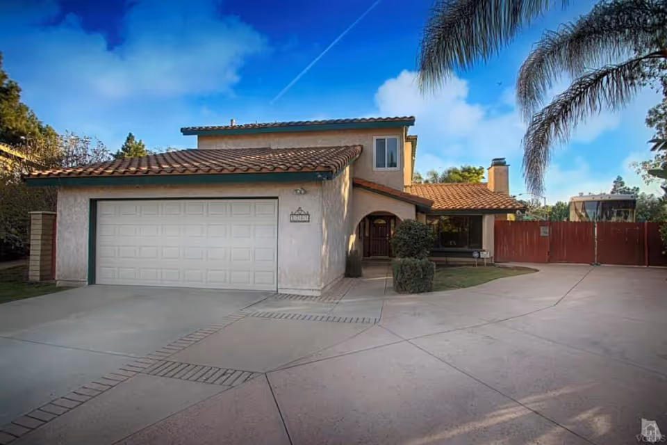 Front exterior view of a two-story house with a tiled roof, a large garage door, a driveway with brick accents, and a red wooden gate. There are palm trees and other greenery around the property under a blue sky with some clouds.