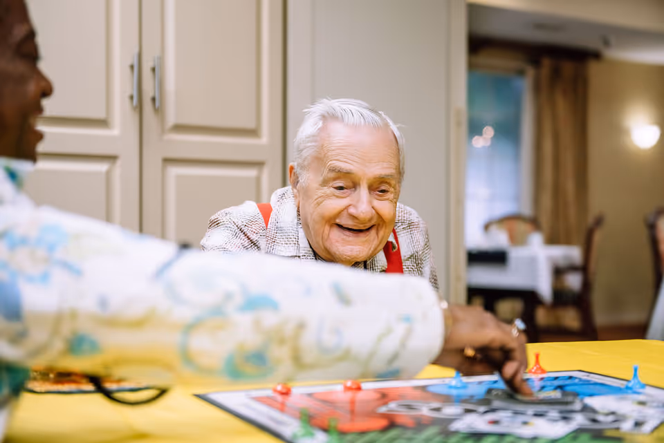 An elderly man with white hair and red suspenders smiling and playing a board game with another person in a well-lit room with beige walls and furniture in the background.
