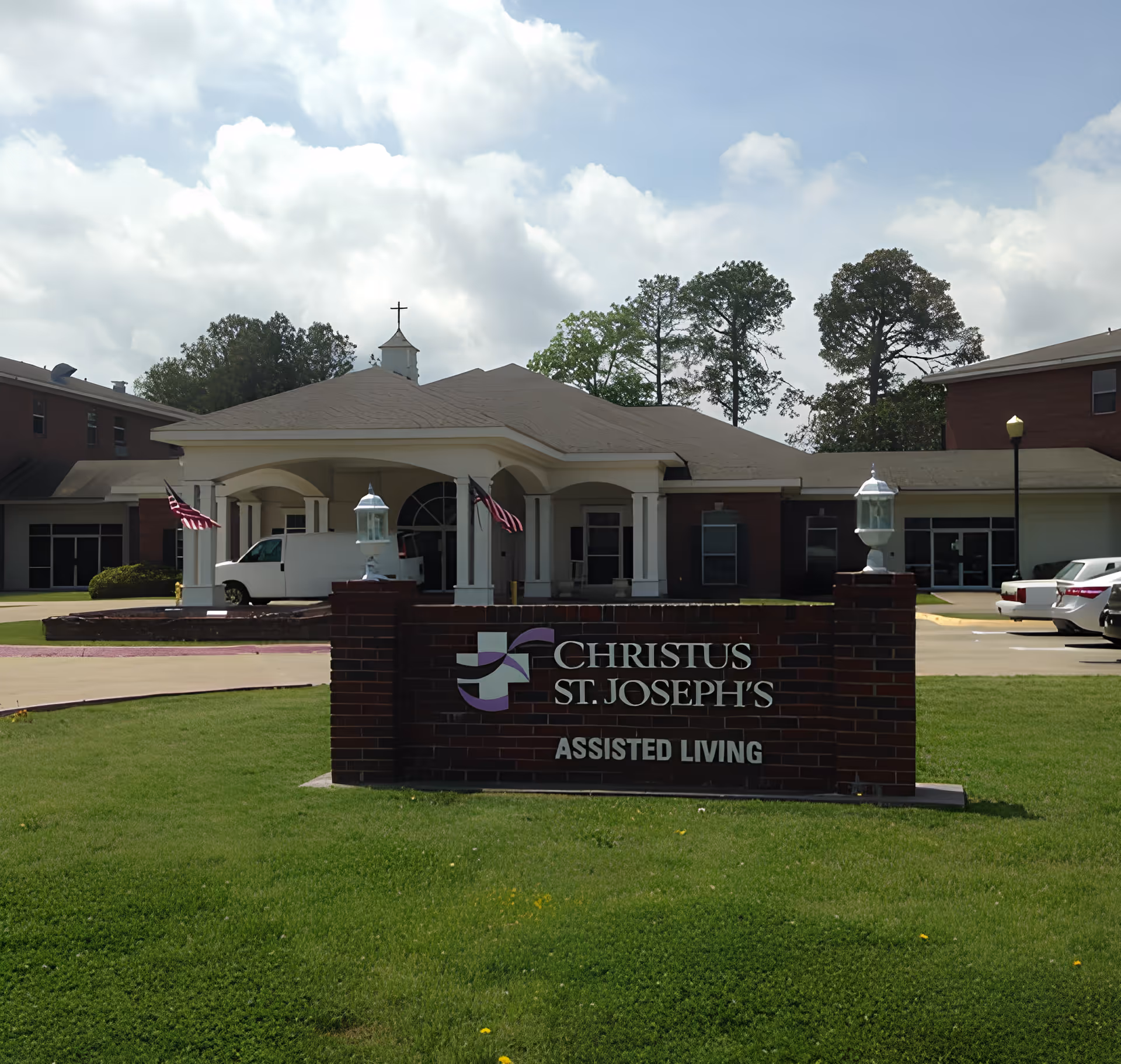 Exterior view of Christus St. Joseph's Assisted Living Center showing the front entrance with a covered driveway, American flags, and a brick sign with the facility's name on a green lawn under a partly cloudy sky.