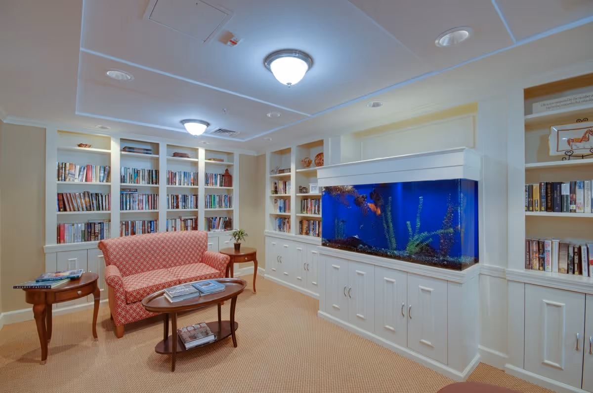 A cozy reading room in Victory Court Senior Apartments featuring built-in white bookshelves filled with books, a large aquarium with colorful fish, a red patterned loveseat, two wooden side tables, and a wooden coffee table with books on it. The room has beige walls and carpet, and ceiling lights providing warm illumination.