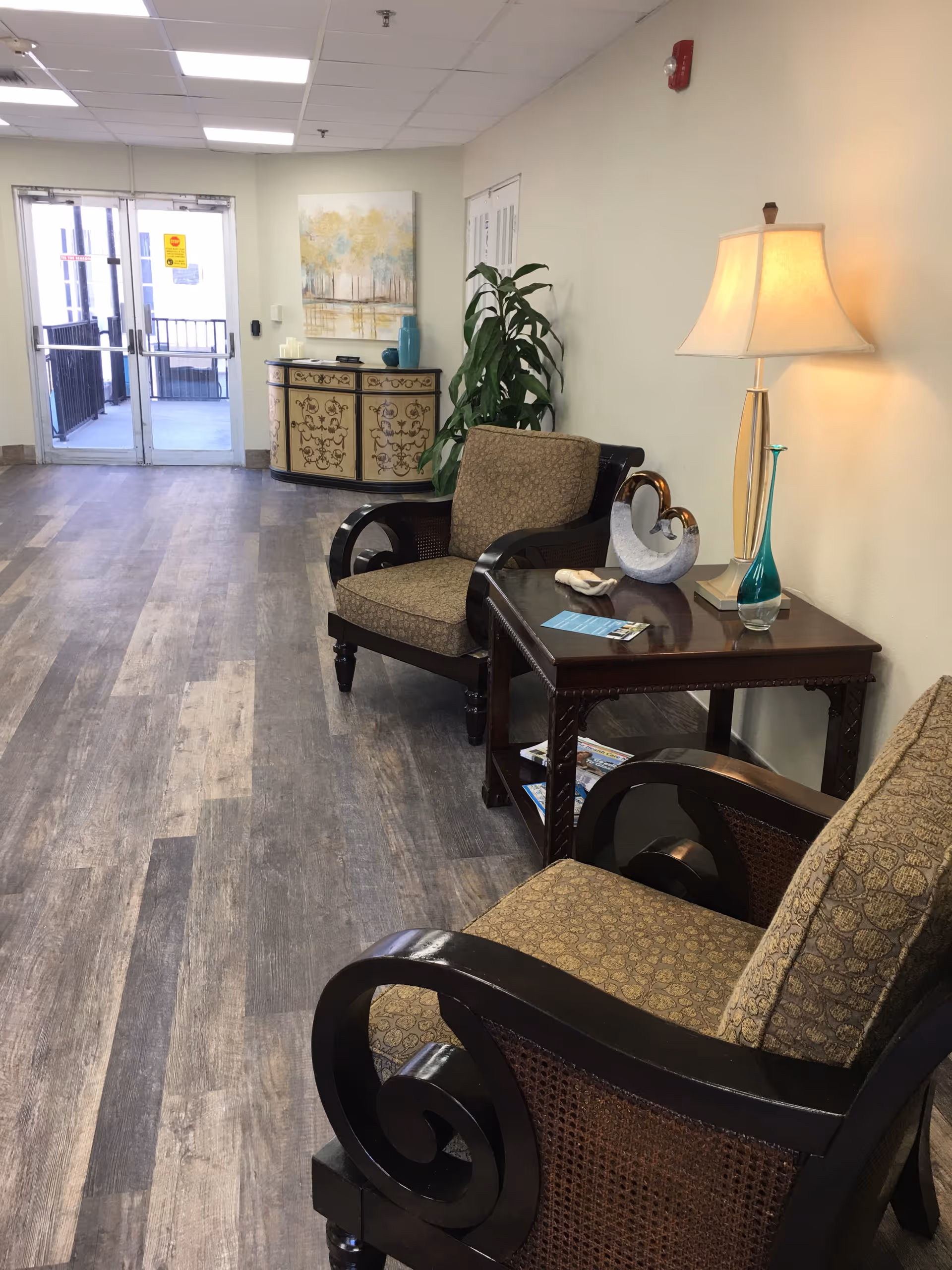 A hallway seating area in a senior living facility with two cushioned armchairs and a wooden side table with a lamp, decorative items, and brochures. The floor has wood-like vinyl planks, and there is a decorative cabinet and a plant against the wall near glass double doors.