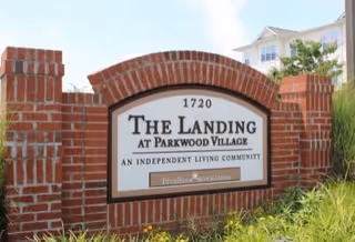 A brick sign at the entrance of The Landing at Parkwood Village, an independent living community, with the address number 1720 displayed above the name. The sign is surrounded by greenery and a building is visible in the background.