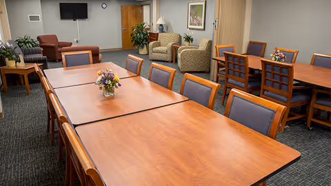 Communal dining and lounge area with wooden tables and chairs, flower centerpieces, armchairs, and a wall-mounted TV.