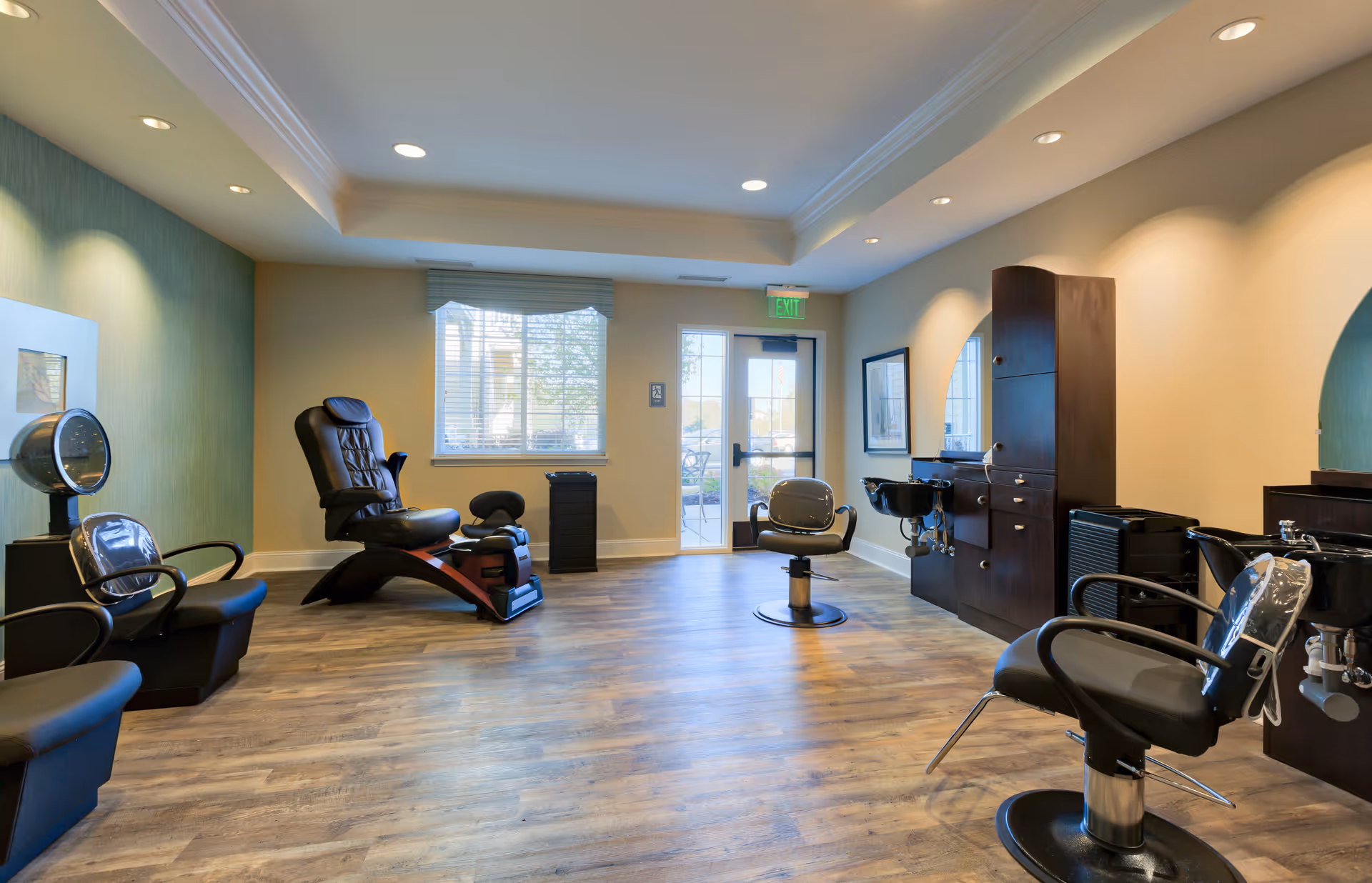 Interior of a senior living facility's hair salon with multiple black salon chairs, hair washing stations, a hair dryer, and a pedicure chair. The room has wooden flooring, recessed ceiling lights, a window with blinds, and a glass door leading outside.