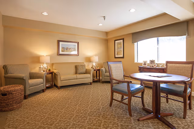A cozy living room with beige walls and carpet, featuring a beige sofa with two cushions, two striped armchairs, a round wooden table with two chairs, two side tables with lamps, framed artwork on the walls, and a window with blinds letting in natural light.