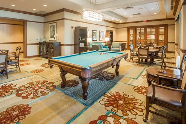 A spacious and well-lit recreational room featuring a pool table in the center, surrounded by multiple seating areas with chairs and tables. The room has a patterned carpet with floral designs, framed artwork on the walls, and warm lighting from ceiling fixtures.