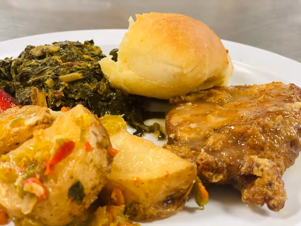 A plate of food featuring cooked greens, a dinner roll, a piece of fried meat with gravy, and seasoned potato wedges.