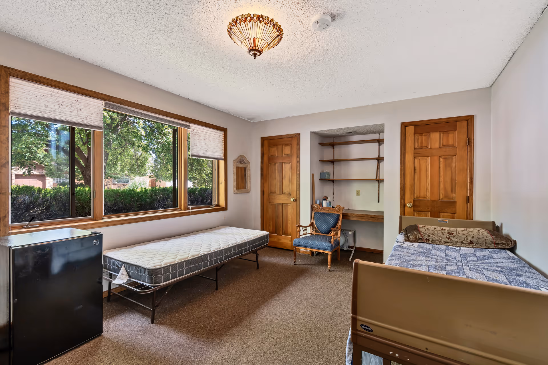 A simple bedroom with two beds, one with a mattress only and the other made with a blue patterned blanket and a decorative pillow. There is a wooden chair with blue upholstery, a small black refrigerator, a built-in desk with shelves, two wooden doors, and a large window showing green trees outside. The ceiling has a decorative light fixture.