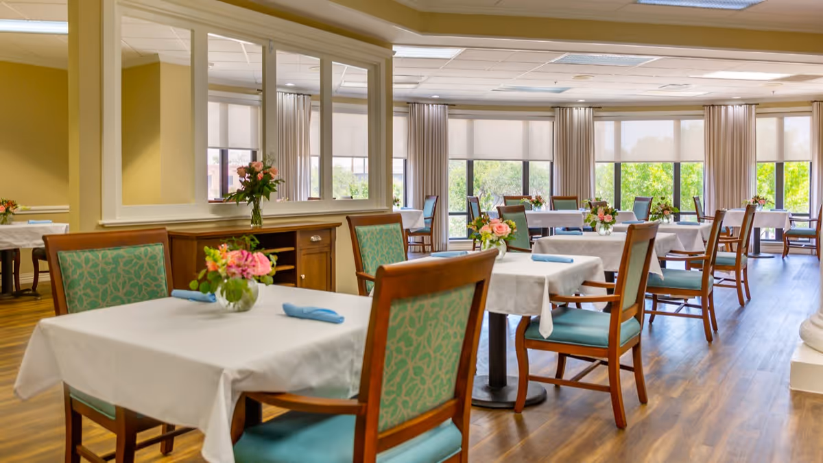 A bright and spacious dining room with multiple tables covered in white tablecloths, each set with a small floral centerpiece and blue napkins. The room features wooden chairs with teal patterned cushions, large windows with beige curtains, and wood flooring.