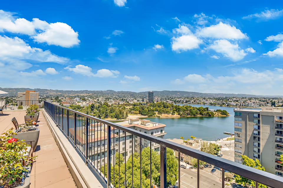 Rooftop balcony with railing and planters overlooking a lake and city skyline under a blue sky.
