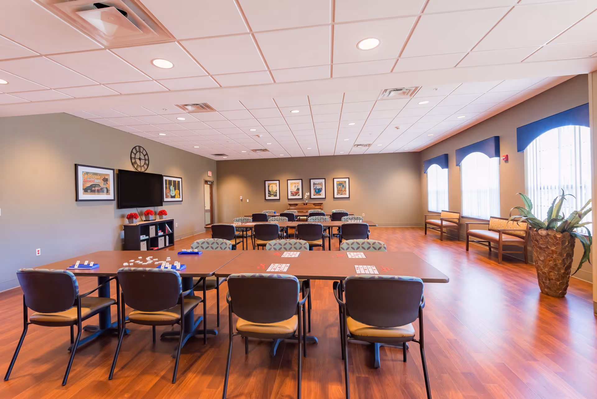 Large bright activity room in a senior living facility with rows of tables and chairs, a TV, and windows.