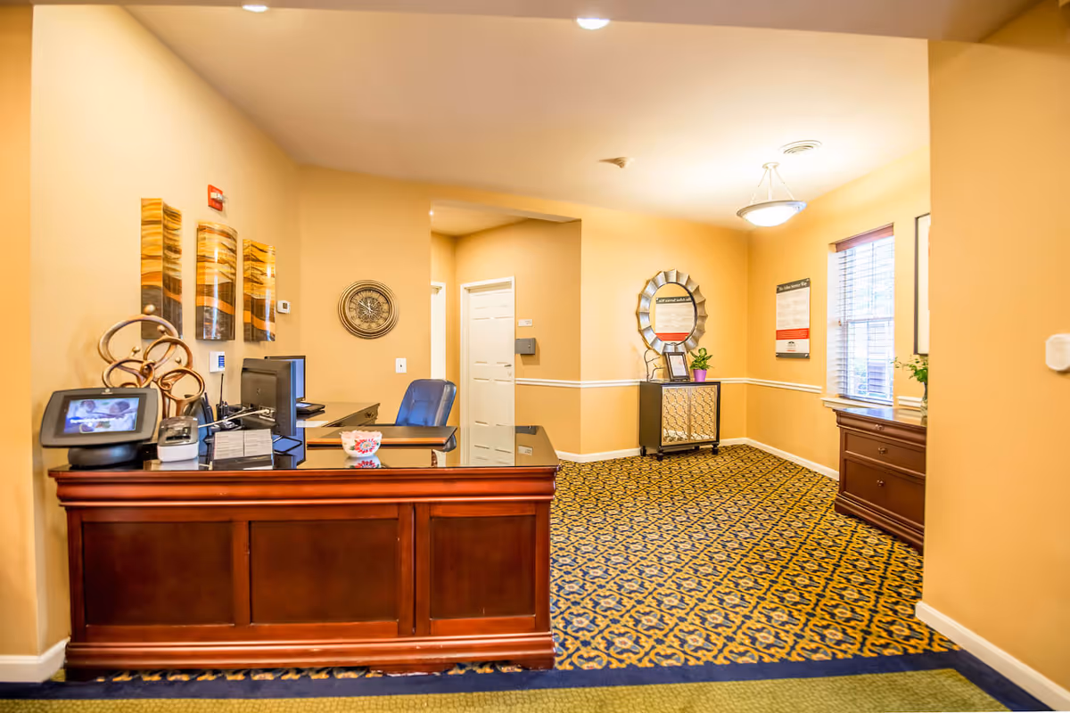 Reception area with a wooden desk, computer, and office chair. The walls are painted beige with decorative wall art and a clock. There is patterned carpet flooring and a small cabinet with a round mirror and a potted plant. A window with blinds lets in natural light.