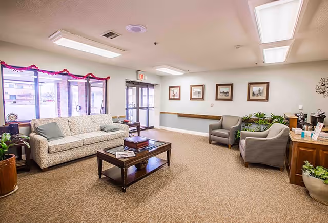 A bright and spacious living room area in a retirement village with a patterned sofa, two armchairs, a wooden coffee table with books and newspapers, framed pictures on the wall, potted plants, and large windows letting in natural light.