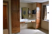 Interior view of a bathroom in a senior living facility featuring a wide sink with a large mirror above it, wooden cabinets on either side, a soap dispenser, and a window with patterned curtains allowing natural light to enter.