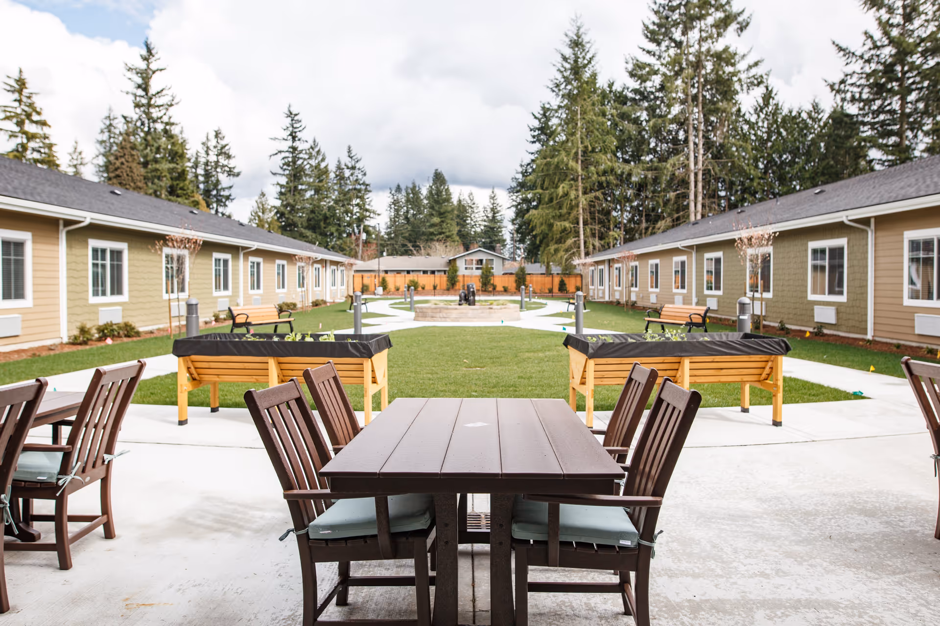 Outdoor courtyard area at Fieldstone Memory Care Marysville featuring wooden tables and chairs on a concrete patio, raised garden beds, green lawn, benches, and surrounded by single-story buildings with trees in the background under a cloudy sky.