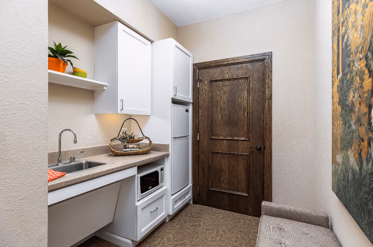 Small kitchenette area with white cabinets, a countertop with a sink, a microwave, and a refrigerator. There is a wooden door at the end of the room, a small bench on the right side, and a painting on the wall above the bench. A small plant and a decorative basket are on the countertop.