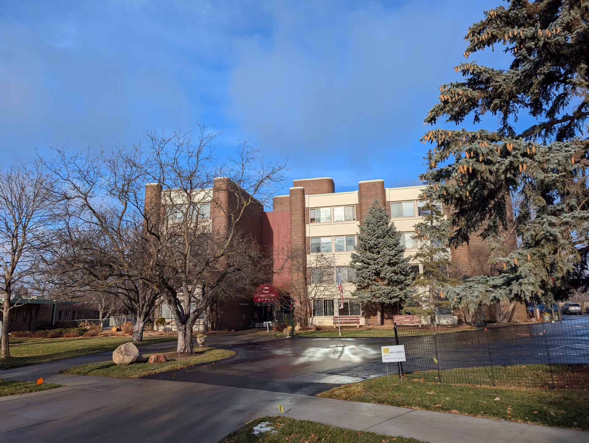 Exterior view of a multi-story brick and beige building with several windows, surrounded by leafless trees and evergreen trees under a partly cloudy blue sky. There is a driveway and a sidewalk leading to the entrance, which has a red awning.