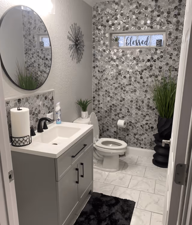 A modern bathroom with a white sink vanity featuring black handles and a black faucet. Above the sink is a round mirror. The wall behind the toilet has a decorative hexagonal tile pattern in shades of gray and white. There is a small window with a 'blessed' sign and small plants on the windowsill. The toilet paper holder is mounted on the wall next to the toilet. The floor is tiled in white with gray veining, and there is a black rug in front of the sink. Green plants in black and white pots add a touch of greenery to the space.