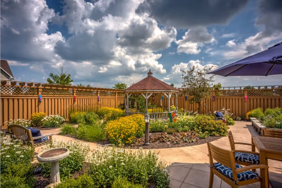 Sunny fenced courtyard garden with a central gazebo, flowering beds, and outdoor seating under an umbrella.