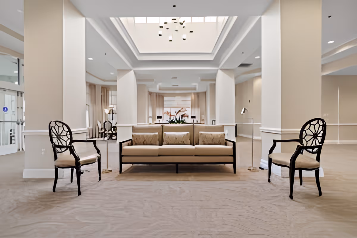 A spacious and well-lit lounge area with a beige sofa and two decorative chairs with black frames and beige cushions. The room features large white columns, a high ceiling with a skylight, and neutral-colored walls and carpet. There are glass doors on the left side and a modern chandelier hanging from the ceiling.