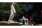 Outdoor garden area featuring a stone statue of a woman in the foreground, with a white gazebo and red umbrellas surrounded by lush green trees in the background under a clear blue sky.