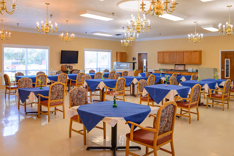 A spacious dining room with multiple tables covered in blue and beige tablecloths, each surrounded by wooden chairs with floral upholstery. The room is well-lit with large windows, several chandeliers hanging from the ceiling, and a television mounted on the wall. Cabinets and a serving area are visible in the background.