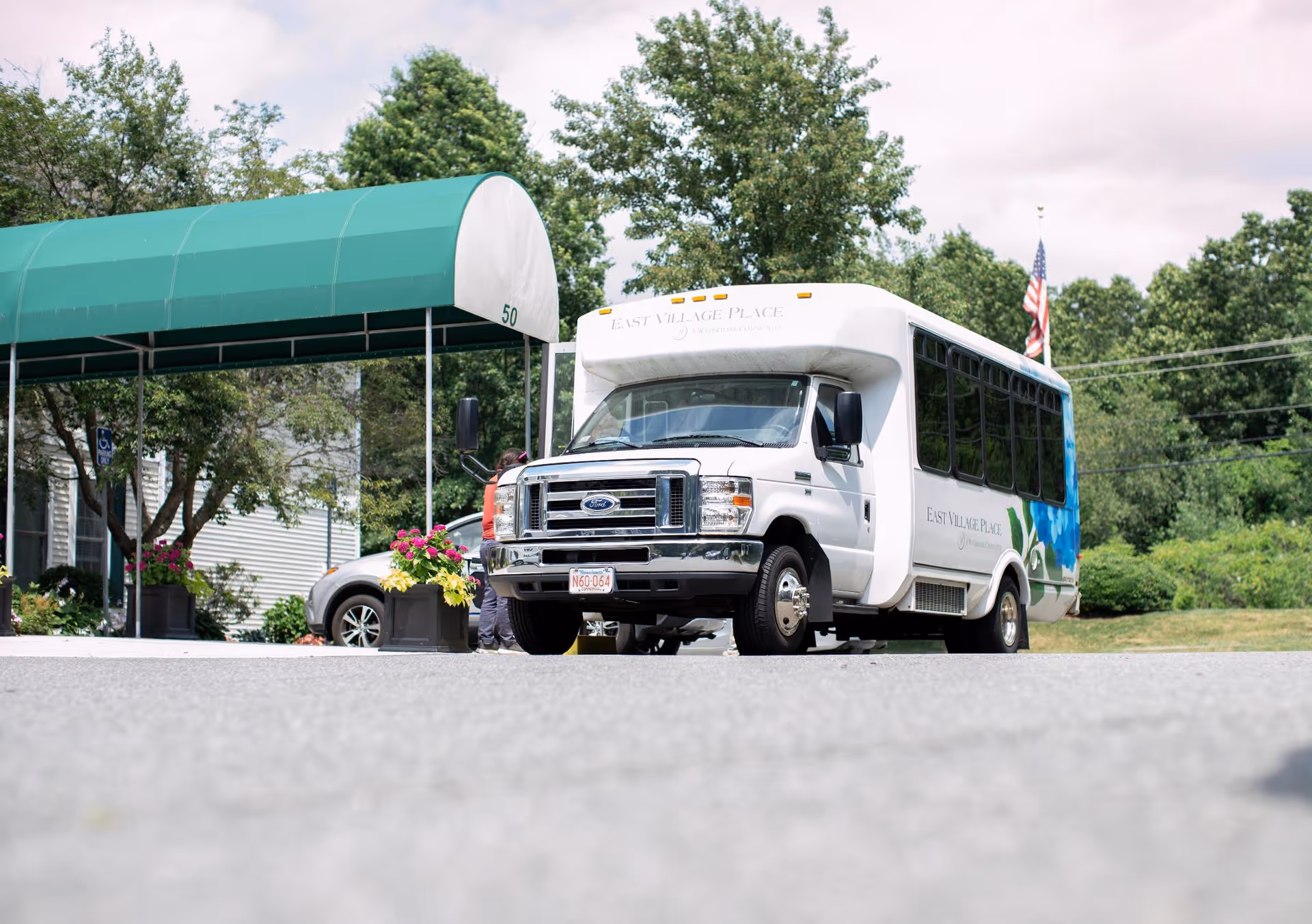 A white shuttle bus with East Village Place branding parked outside a building entrance with a green canopy. There are trees and plants around, and an American flag is visible in the background.