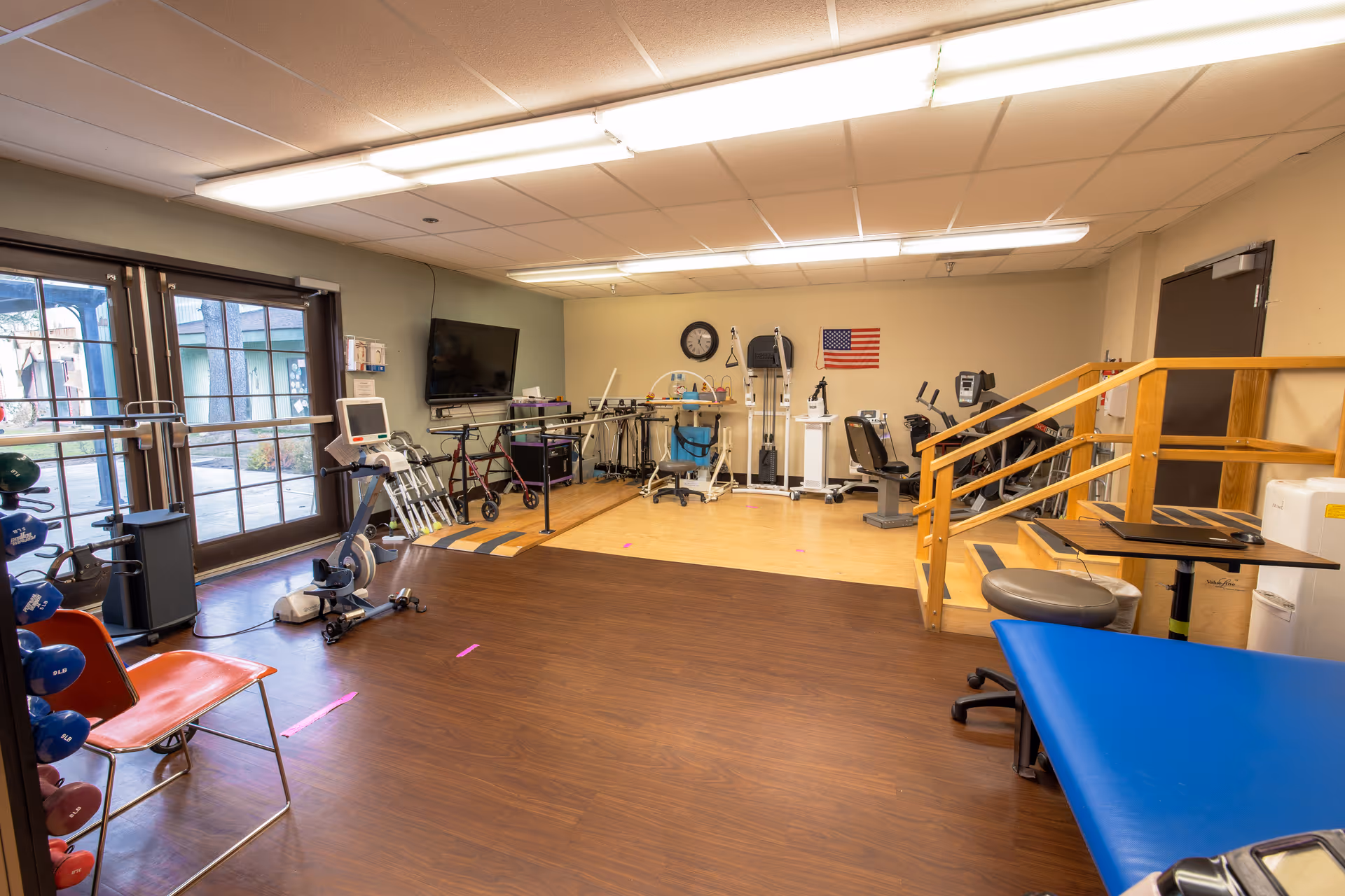 A rehabilitation or physical therapy room with exercise equipment including stationary bikes, parallel bars, weights, and various therapy devices. There is a large windowed door on the left side, a clock and an American flag on the far wall, and wooden handrails with steps on the right side.