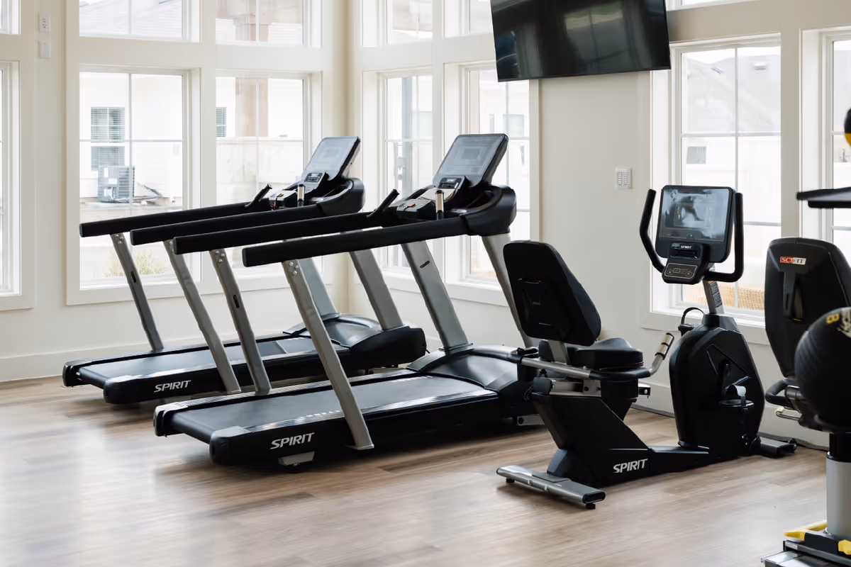 A bright fitness room with large windows showing outdoor views. The room contains two Spirit treadmills, a recumbent exercise bike, and an upright exercise bike, all positioned on a light wood floor. A large flat-screen TV is mounted on the wall above the exercise bikes.