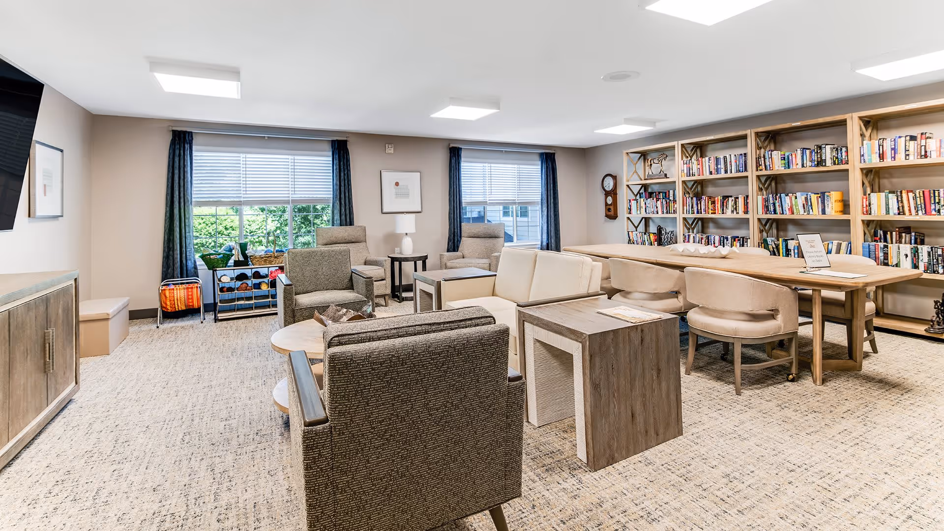 A bright and spacious common area in a senior living facility with comfortable seating including armchairs and sofas arranged around coffee tables. Large windows with blue curtains let in natural light. A long wooden table with chairs is positioned near a wall filled with bookshelves stocked with books and decorative items. The room has a neutral color palette with beige walls and carpeted flooring.