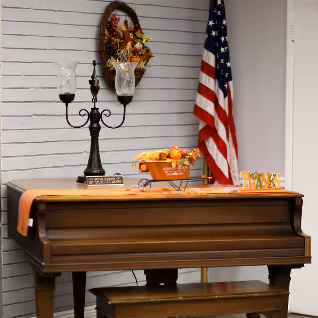 A wooden piano with a matching bench in front of a gray brick wall. On top of the piano are a black candle holder with two glass candle covers, a small decorative wheelbarrow filled with autumn-themed decorations, and a small sign spelling 'THANKS'. An American flag stands in the corner behind the piano, and a fall-themed wreath hangs on the wall above.