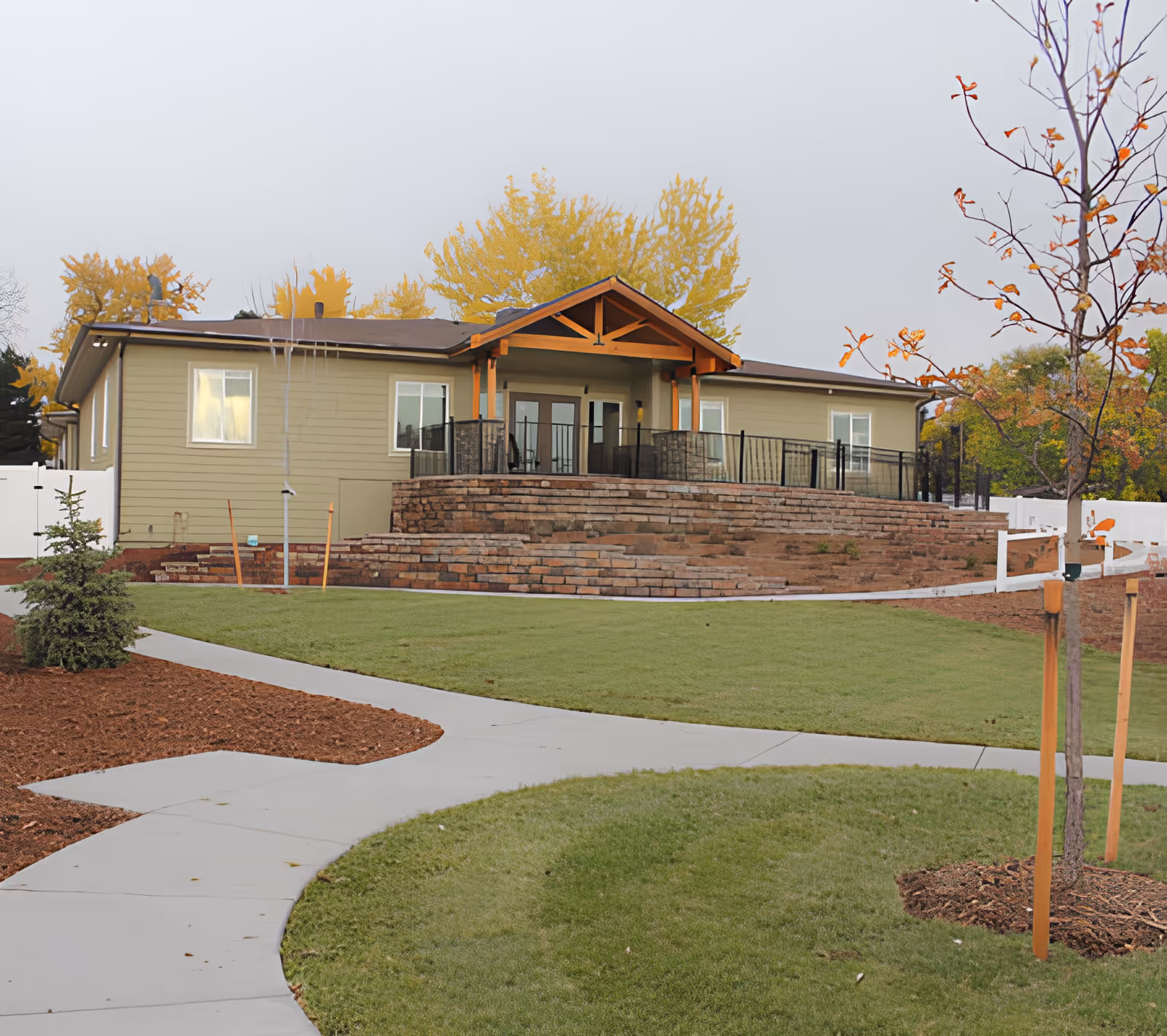 Exterior view of a single-story assisted living facility building with beige siding and a brown roof. The building features a covered entrance with wooden beams and a stone retaining wall in front. There is a curved concrete walkway leading up to the building, surrounded by green grass, newly planted trees, and mulch beds. Trees with autumn foliage are visible in the background under a cloudy sky.