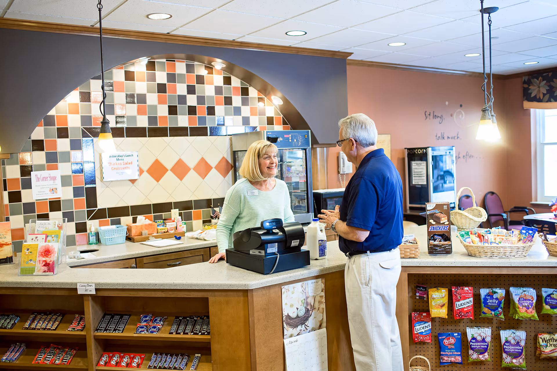 A woman behind a counter smiling and talking to an older man in a casual setting that appears to be a small shop or snack bar inside a senior living facility. The counter has a cash register, snacks, and candy displayed. The background features a tiled wall with a menu and various items on shelves.