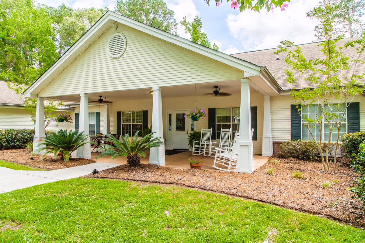 Front porch of a single-story building with white siding and green shutters, featuring white rocking chairs, hanging flower baskets, ceiling fans, and landscaped greenery including small palm plants and bushes.