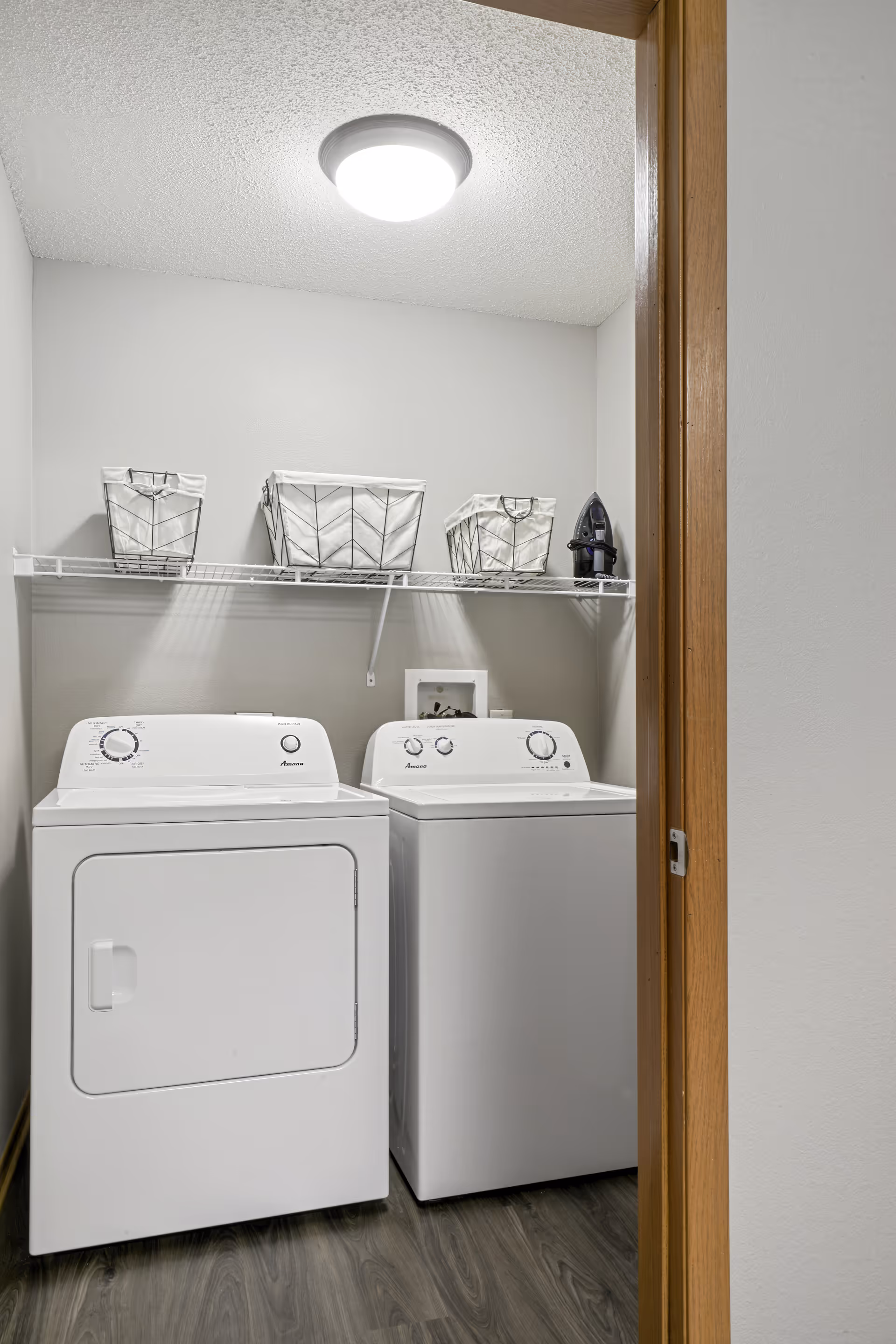 Small laundry room with a white washer and dryer under a wire shelf holding baskets and an iron.