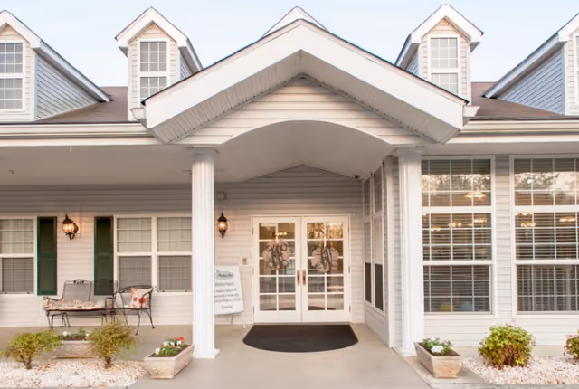Front entrance of a senior living facility with white double doors, a covered porch supported by two white columns, and windows with white shutters. There is a bench with cushions on the left side and potted plants on both sides of the entrance.
