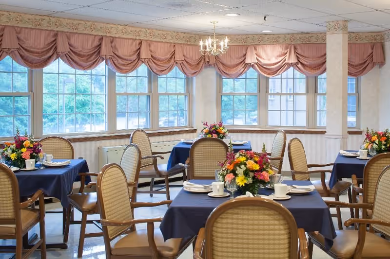 Bright dining room with several tables covered in blue tablecloths, floral centerpieces, place settings, and chairs near large windows.