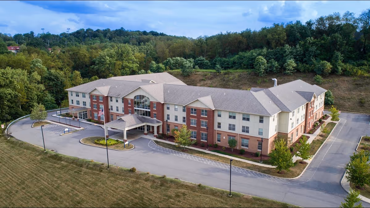Aerial view of a large three-story senior living facility building with a covered entrance, surrounded by a driveway and parking areas. The building has a combination of brick and light-colored siding exterior, with multiple windows and a wooded area in the background.