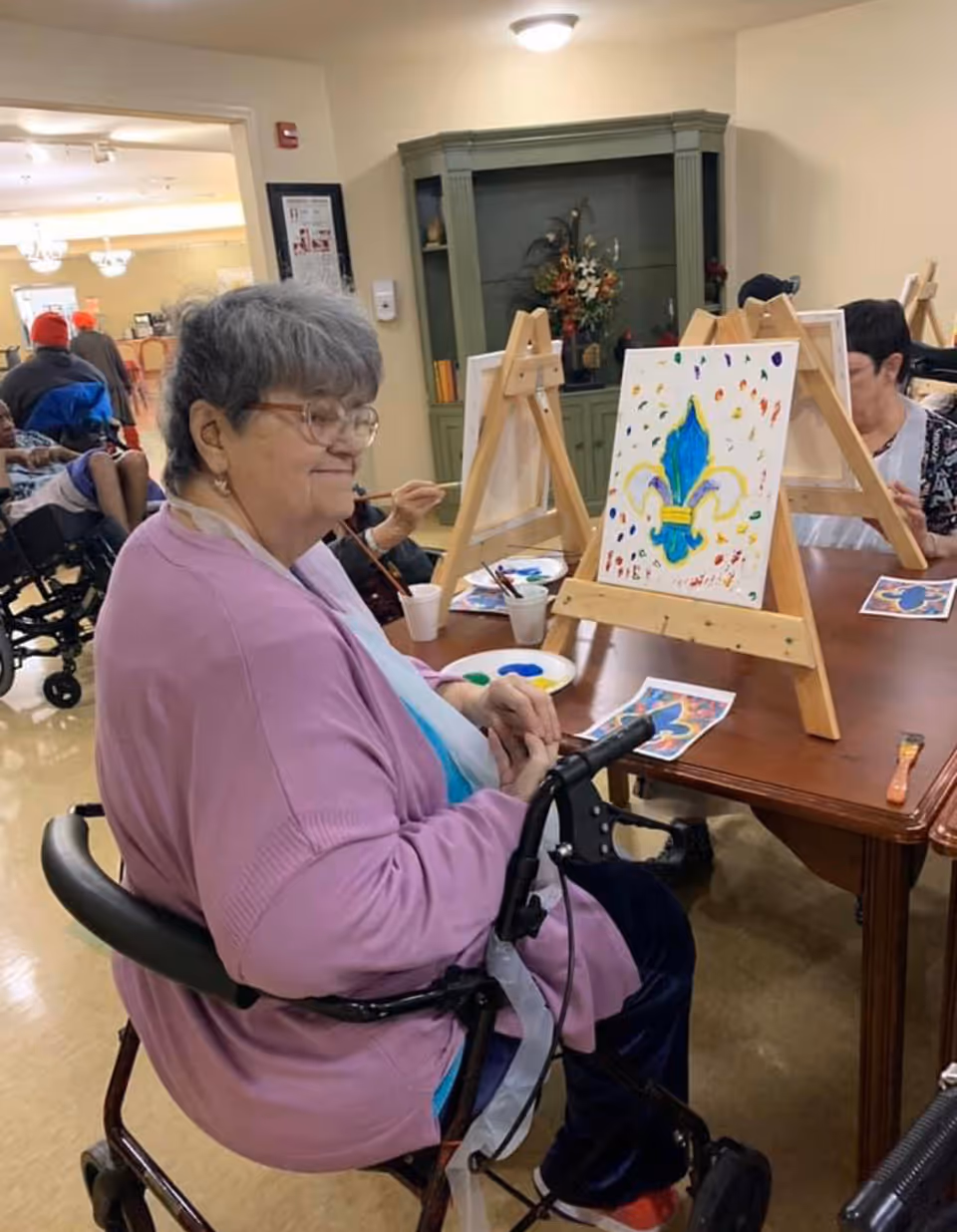 An elderly woman in a wheelchair wearing a pink cardigan and glasses is seated at a table painting on a canvas placed on an easel. The canvas shows a colorful fleur-de-lis design. Other people are also seated at the table painting. The room has a cabinet with decorative items and a bright, welcoming atmosphere.