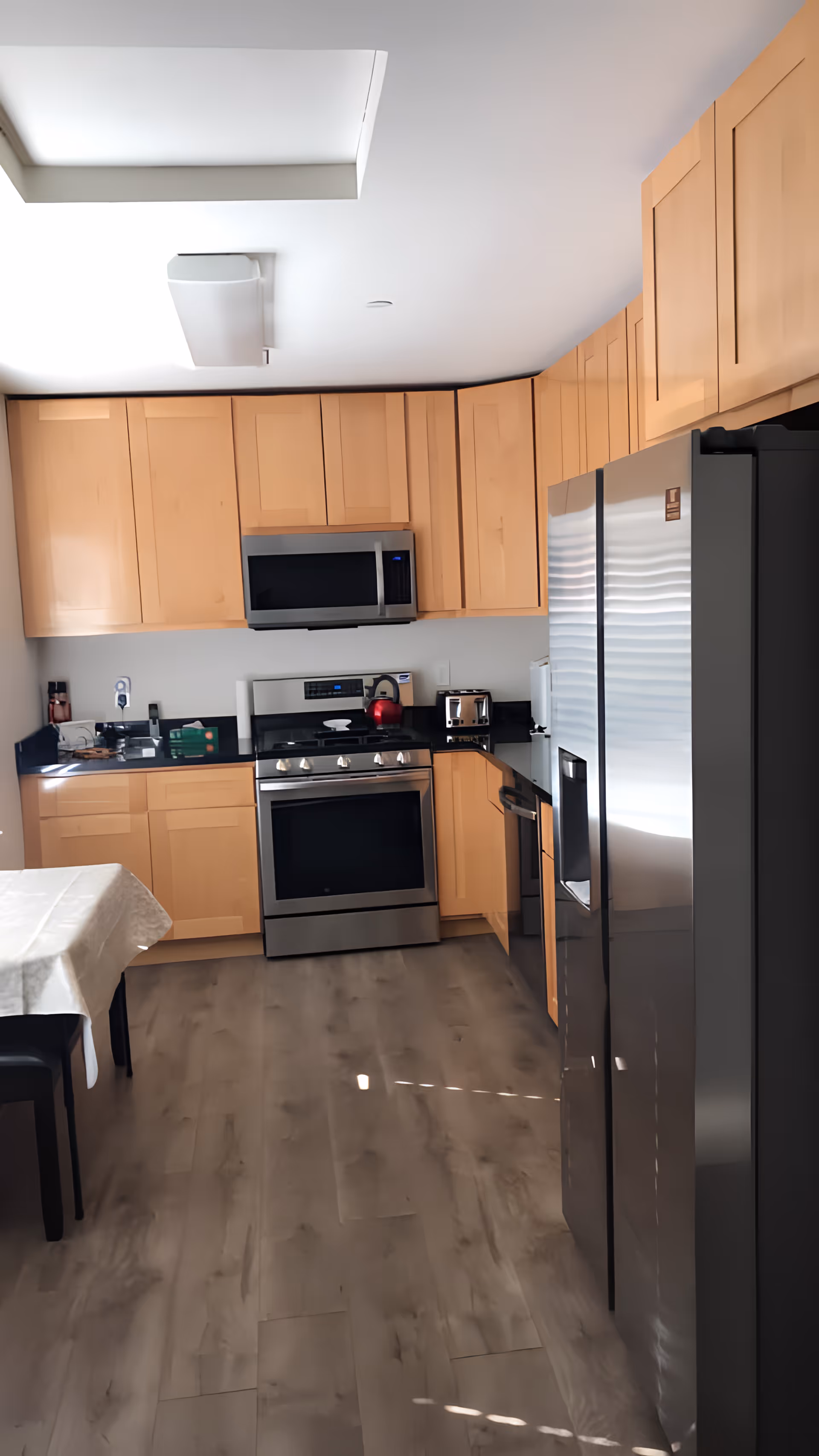 Modern kitchen with light wood cabinets, stainless steel appliances including a refrigerator, oven, microwave, and dishwasher. There is a black countertop with a red kettle and toaster on it. A dining table with a white tablecloth and black chairs is partially visible on the left side. The floor is a light wood laminate.