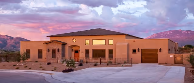 Single-story building with a beige exterior and a brown roof, set against a backdrop of mountains and a colorful sunset sky. The building has multiple windows, a front door with a small porch, and a driveway leading to a garage on the right side. Desert landscaping with rocks and sparse vegetation surrounds the building.