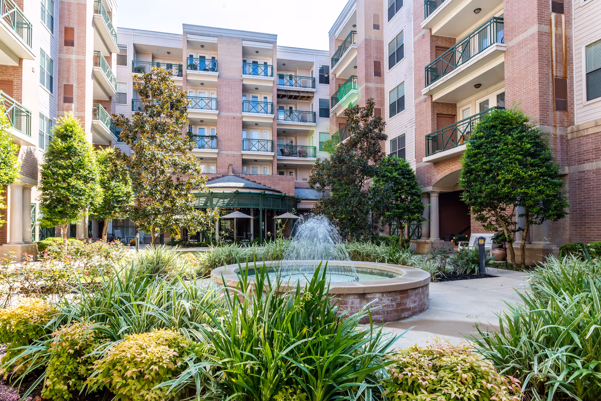 Outdoor courtyard area of a senior living facility with a central circular water fountain surrounded by lush green plants and trees, multi-story brick and beige buildings with balconies in the background, and a covered seating area with umbrellas.