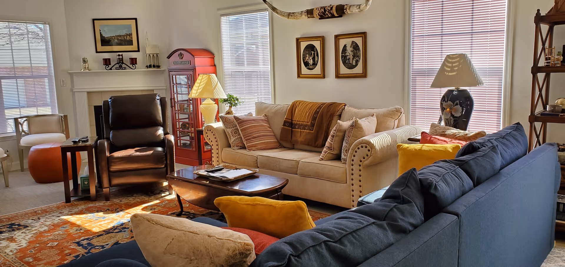 A cozy living room with a beige couch adorned with multiple pillows and a throw blanket, a dark blue couch with colorful pillows, a brown leather recliner, a white armchair, and a wooden coffee table. The room features a fireplace with decorative items on the mantel, a red vintage-style phone booth, two windows with blinds, framed pictures on the walls, a large decorative horn mounted above the couch, and two table lamps providing warm lighting.