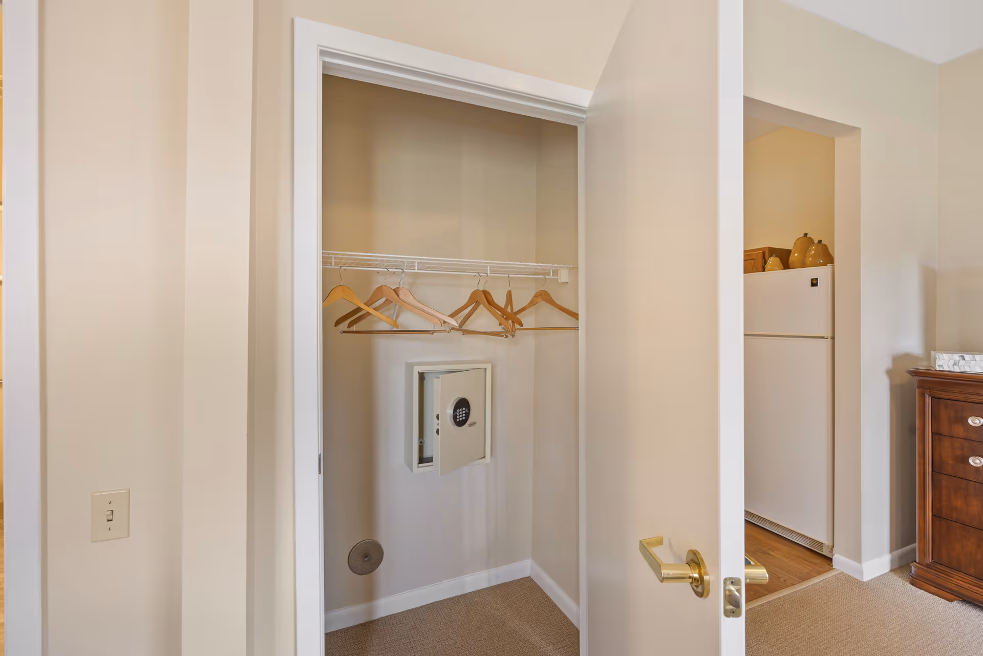 Open closet with several wooden hangers on a wire shelf and a small open safe mounted on the back wall. The closet door is partially open, revealing part of an adjacent room with a white refrigerator and a wooden dresser.