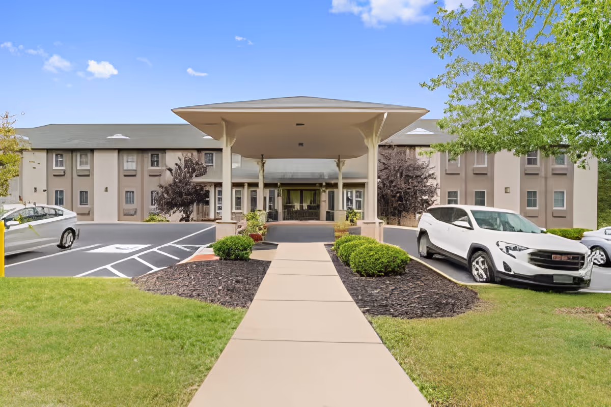 Front exterior view of Rittenhouse Village At Muhlenberg, showing a covered entrance with a walkway leading up to it, surrounded by landscaped bushes and grass, with parked cars on either side and a clear blue sky above.