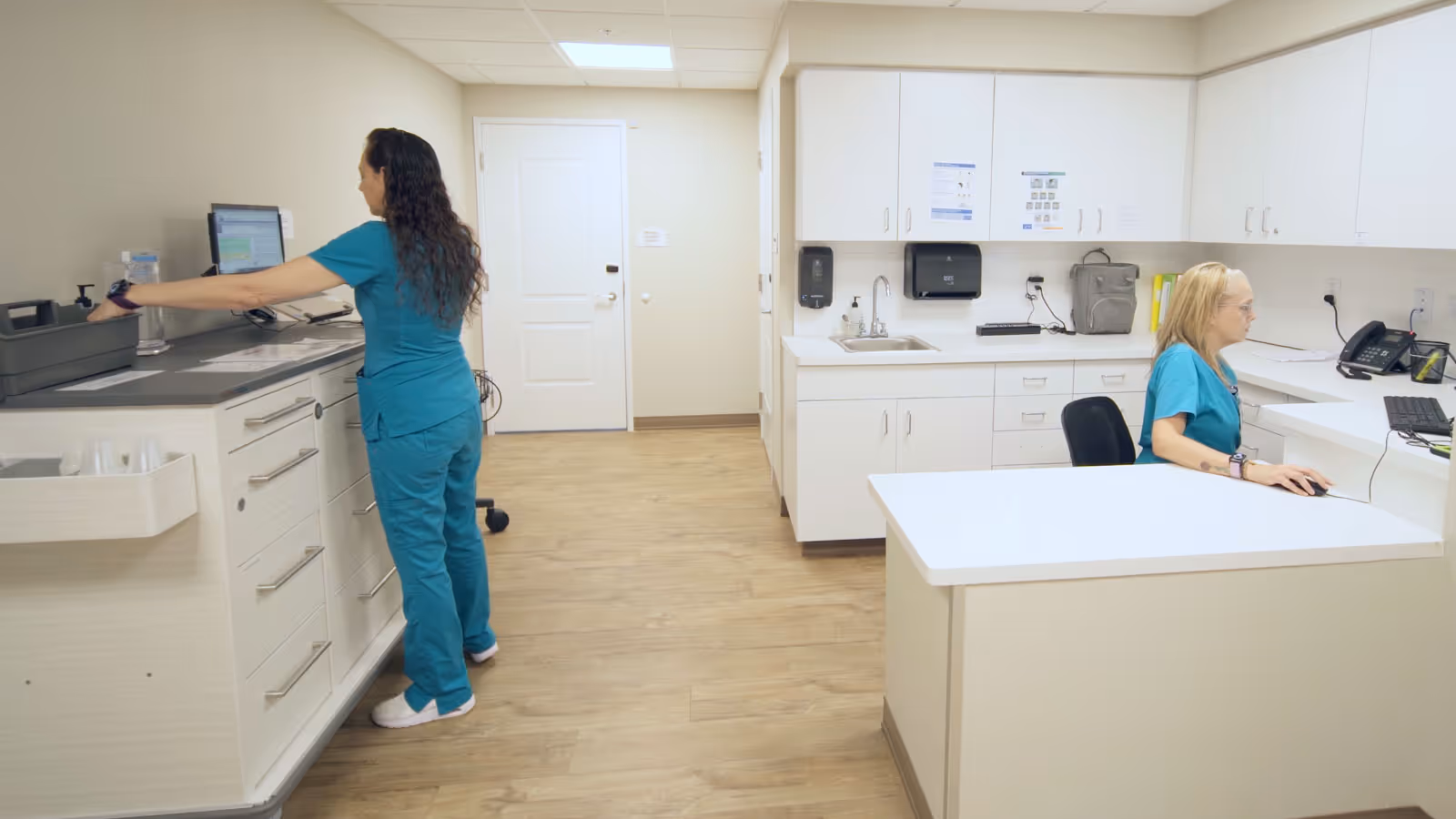Two healthcare workers in teal scrubs are working in a clean, well-lit medical or administrative office area. One is standing and using a computer on a counter with drawers, while the other is seated at a desk working on a computer. The room has white cabinets, a sink, and light wood flooring.