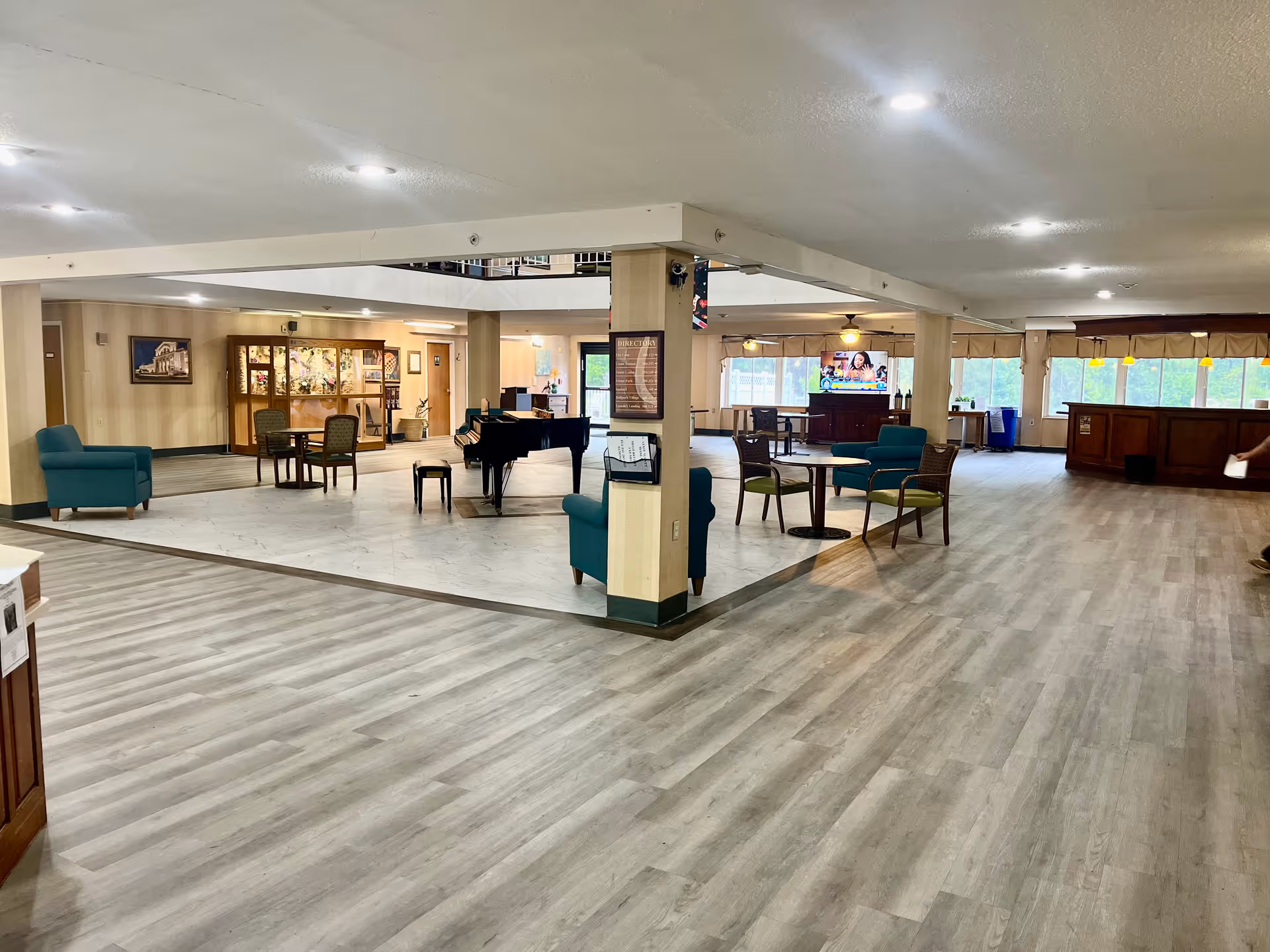 Spacious interior common area of a senior living facility with light wood flooring, several seating arrangements including armchairs and small tables, a black grand piano in the center, a television mounted on a stand, and large windows letting in natural light.
