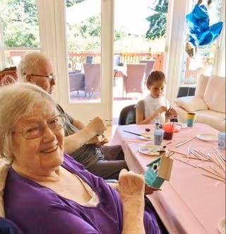 An elderly woman smiling at the camera while holding a small craft project at a table covered with a pink tablecloth. Behind her, an elderly man and a young girl are also engaged in crafting activities. The room has large glass doors leading to an outdoor patio with chairs and greenery visible outside.